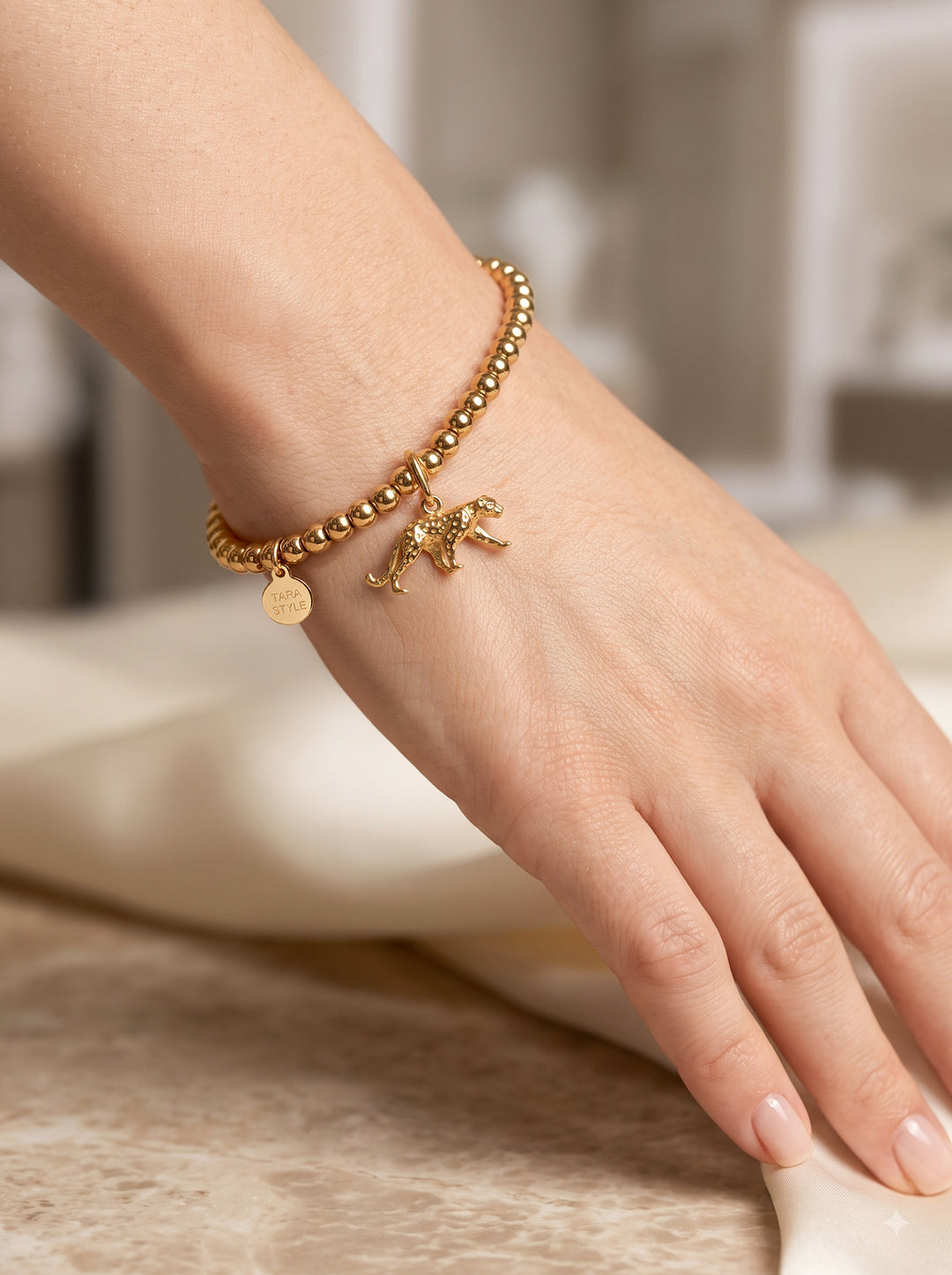 Nahaufnahme eines weiblichen Handgelenks, das ein goldenes Perlenarmband trägt. Zwei goldene Anhänger hängen herab: ein detaillierter, gehender Jaguar und eine kleine, runde Plakette mit der eingravierten Aufschrift "TARA STYLE". Die Hand liegt entspannt auf einer cremefarbenen, fließenden Stoffbahn über einer Marmoroberfläche.