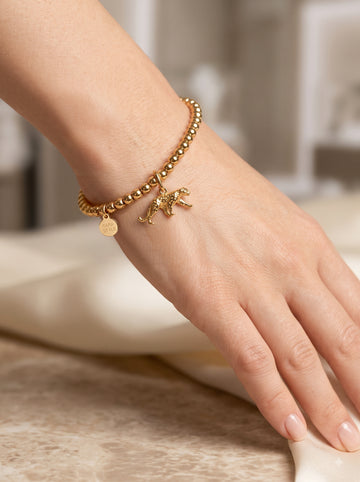 Nahaufnahme eines weiblichen Handgelenks, das ein goldenes Perlenarmband trägt. Zwei goldene Anhänger hängen herab: ein detaillierter, gehender Jaguar und eine kleine, runde Plakette mit der eingravierten Aufschrift "TARA STYLE". Die Hand liegt entspannt auf einer cremefarbenen, fließenden Stoffbahn über einer Marmoroberfläche.