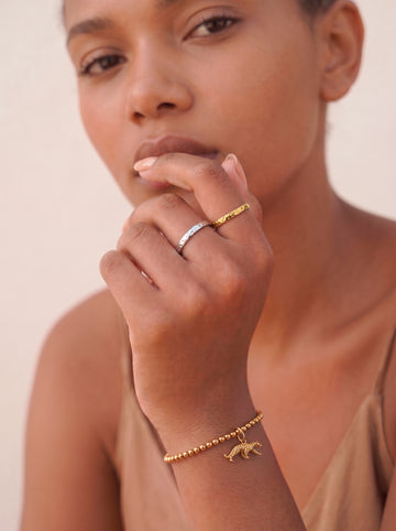Nahaufnahme eines weiblichen Handgelenks, das ein goldenes Perlenarmband trägt. Zwei goldene Anhänger hängen herab: ein detaillierter, gehender Jaguar und eine kleine, runde Plakette mit der eingravierten Aufschrift "TARA STYLE". Die Hand liegt entspannt auf einer cremefarbenen, fließenden Stoffbahn über einer Marmoroberfläche.